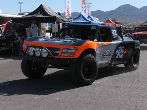 1990 Ford Bronco - orange and gray off-road race truck numbered 22 with roof lights and K&N sponsorship driving through a desert pit area.