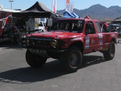 1990 Ford Bronco - red off-road race truck numbered 758 with Goodyear and sponsor decals, a brush guard, and yellow lights in a busy pit area.
