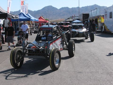 1990 Ford Bronco - silver open tube-frame off-road race buggy numbered 1605 rolling through a crowded desert pit with spectators and tents.