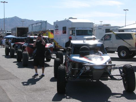 1990 Ford Bronco - Several off-road race buggies and trucks staged among motorhomes and trailers in a sunny paved pit area with people walking.