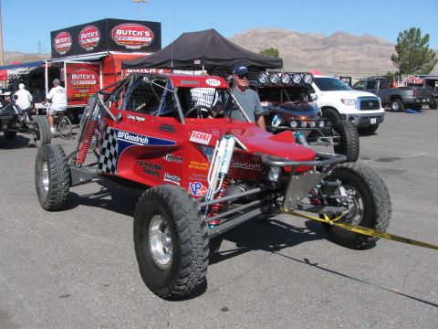 1990 Ford Bronco - red BFGoodrich-decaled open tube-frame off-road race buggy strapped to the pavement with a man standing beside it.