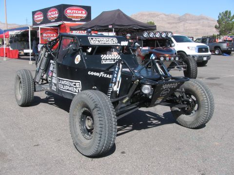 1990 Ford Bronco - black open tube-frame off-road race buggy with Lawrence Equipment and Goodyear decals parked on pavement near pit canopies.