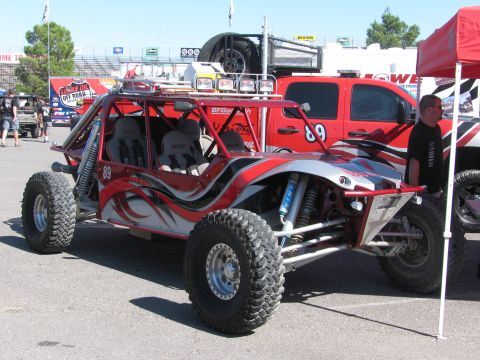 1990 Ford Bronco - red and silver four-seat open off-road race buggy numbered 89 with a roof-mounted spare tire and lights in a pit area.