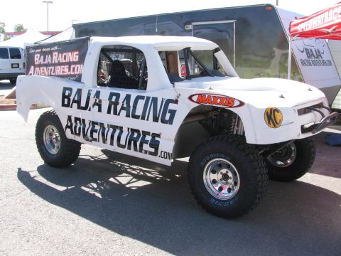 1990 Ford Bronco - white off-road race truck lettered Baja Racing Adventures with KC and Maxxis decals parked beside a trailer.