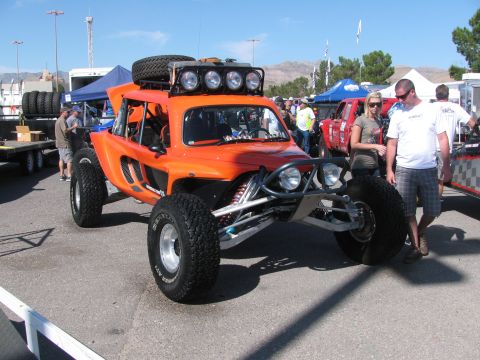 1990 Ford Bronco - orange-bodied open off-road race buggy with a roof-mounted spare and light bar on display as spectators look on in a pit.