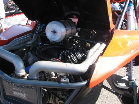 1990 Ford Bronco - close-up of an off-road race vehicle's engine bay showing the motor, exhaust headers, air filter, and shock under an orange body panel.