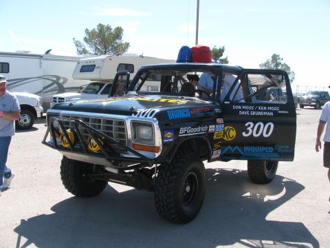 1990 Ford Bronco - black classic Ford Bronco race truck numbered 300 with a brush guard, yellow KC lights, and sponsor decals in a desert pit.