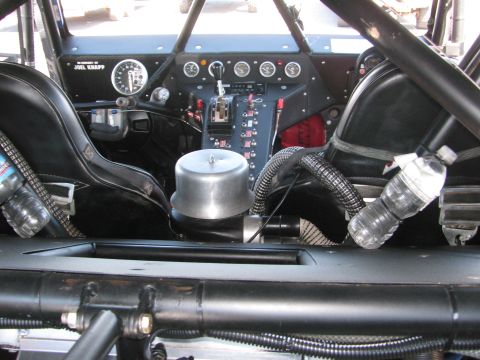 1990 Ford Bronco - Interior of a tube-chassis off-road race vehicle showing two black seats, a center gauge panel, shifter and zip-tied water bottles.