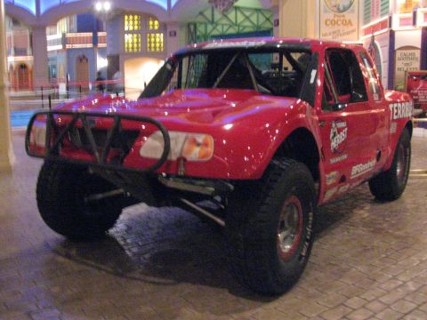 1990 Ford Bronco - Front three-quarter view of a red Terrible Herbst off-road trophy truck with a tubular front bumper on display inside a venue.