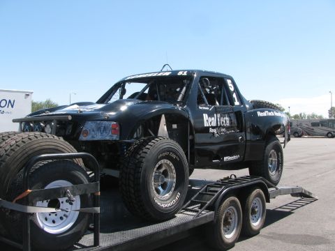 1990 Ford Bronco - black fiberglass-bodied off-road race truck numbered 51 with RealTech Realty decals sits on a tandem-axle flatbed trailer.