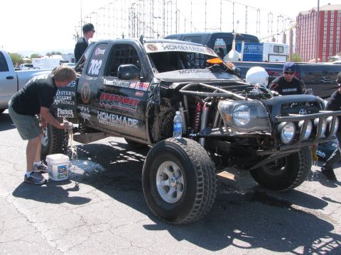 1990 Ford Bronco - Crew members wash a black number 702 Homemade Motorsports off-road race truck with a Toyota grille in a paved paddock.