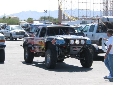 1990 Ford Bronco - black off-road race truck with orange flame graphics and a tube front bumper drives through a paved paddock with spectators and tow rigs.