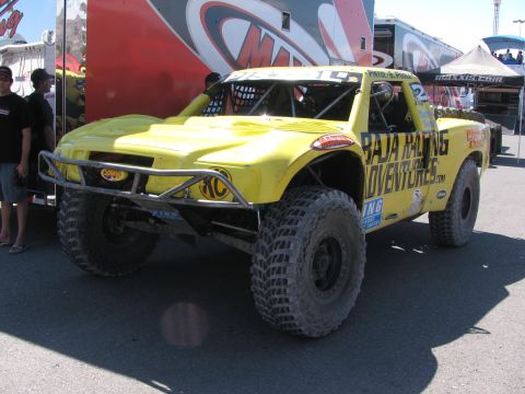 1990 Ford Bronco - yellow off-road race truck with Baja Racing Adventures decals and a tube front bumper parked in a paddock near a Maxxis-branded trailer.