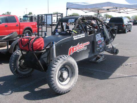 1990 Ford Bronco - black tube-frame off-road race buggy numbered 1606 with Robinson Racing decals and a red gear bag parked under a canopy in a paddock.