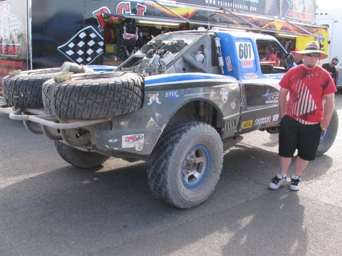 1990 Ford Bronco - young person stands beside a gray and blue number 601 off-road race truck with spare tires up front, parked near a PCI race radios trailer.