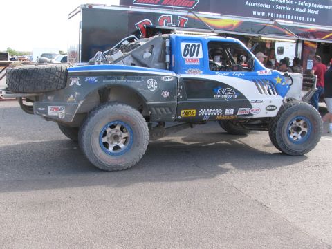 1990 Ford Bronco - Front three-quarter view of a blue and gray number 601 RPB Motorsports off-road race truck with blue beadlock wheels in a paddock.