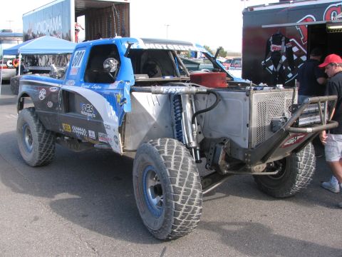 1990 Ford Bronco - Rear three-quarter view of a blue and gray number 601 off-road race truck showing the cut-out bed, rear shocks and crew working nearby.