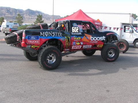 1990 Ford Bronco - Rear three-quarter view of a number 602 off-road trophy truck wrapped in Monster Energy, Boost Mobile and Ford graphics in a desert paddock.