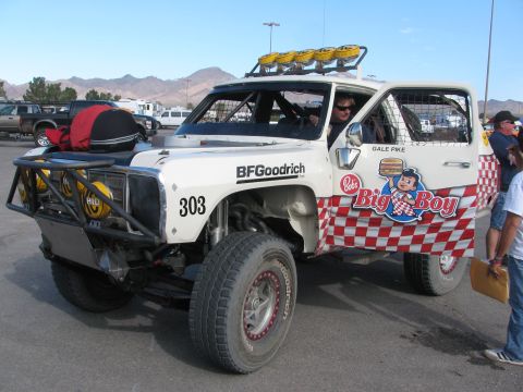 1990 Ford Bronco - white tube-frame off-road race truck numbered 303 wears Big Boy checkered livery and BFGoodrich decals in a desert paddock.