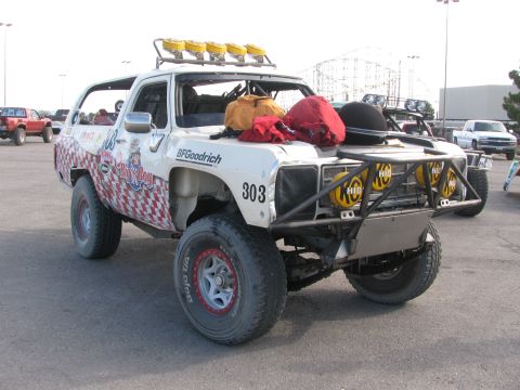 1990 Ford Bronco - 303 off-road race truck sits in a paved lot with a roof-mounted light bar and amusement park rides visible behind it.