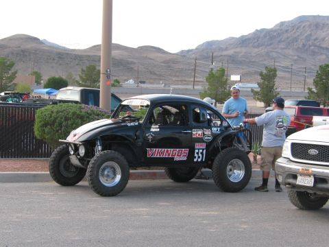 1990 Ford Bronco - black VW Baja Bug race car numbered 551 with Vikingos livery sits in a lot while two men stand alongside it.