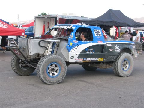 1990 Ford Bronco - blue and white tube-frame off-road race truck numbered 601 with a helmeted driver idles in a busy race paddock.