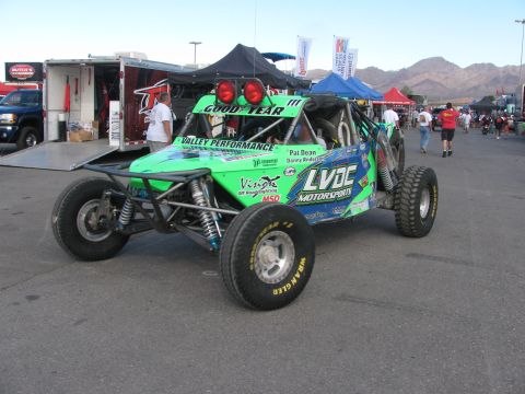 1990 Ford Bronco - green tube-frame off-road racing buggy lettered LVDC Motorsports and Goodyear sits among trailers and canopies.