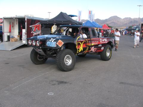 1990 Ford Bronco - black desert race truck with flame graphics and a four-pod light bar parked beside trailers and pop-up tents.
