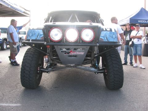 1990 Ford Bronco - Front-on view of a tube-frame off-road race car showing three round driving lights and exposed suspension as spectators watch.