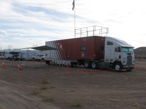 1990 Ford Bronco - maroon cabover semi tractor-trailer serving as a race support rig sits in a dirt lot beneath an American flag and awning.