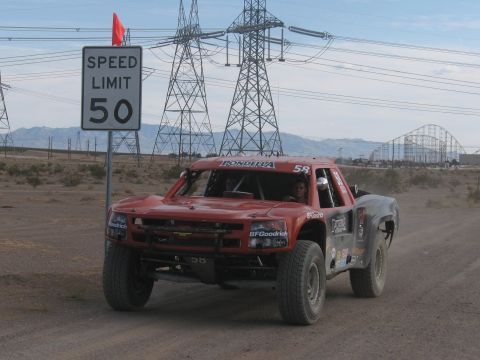 1990 Ford Bronco - weathered red off-road race truck drives along a dirt course past a Speed Limit 50 sign with power lines overhead.