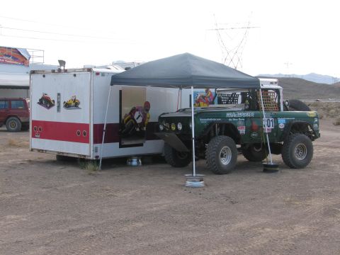 1990 Ford Bronco - green off-road race vehicle numbered 301 parked under a canopy next to an open enclosed trailer in a dusty staging area.
