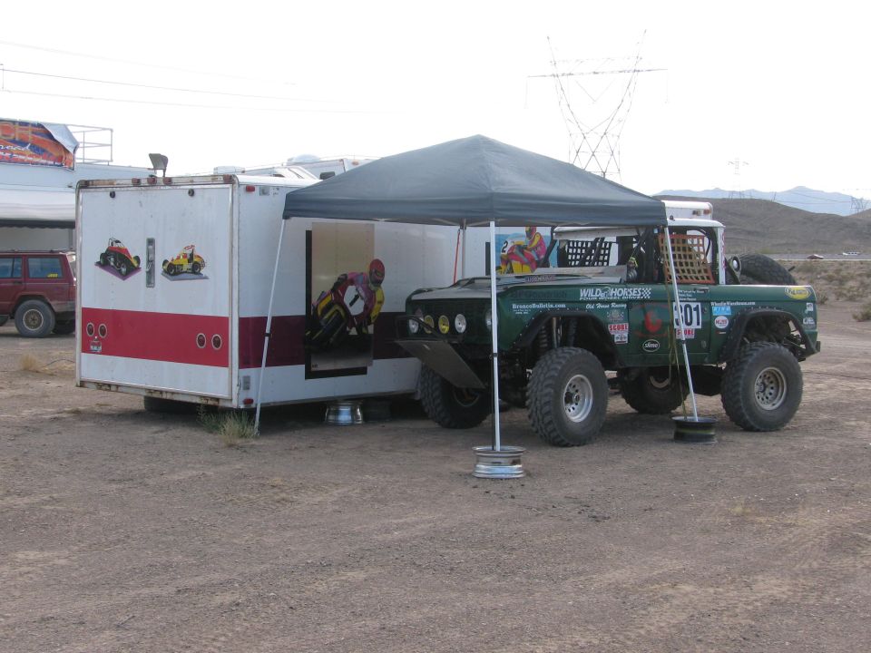 Green off-road racer #301 under a canopy beside an enclosed trailer in the desert