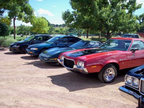 1972 Ford Ranchero - A row of cars are parked on a gravel lot under trees on a sunny day.