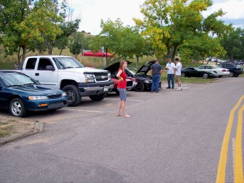 1972 Ford Ranchero - A group of people stand around several cars parked in a lot, with a white pickup truck and a teal sedan in the foreground.