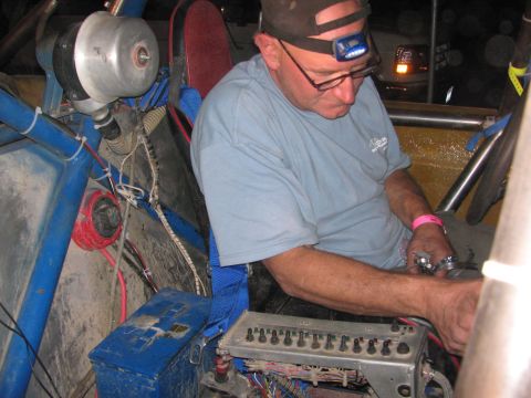 1990 Ford Bronco - man wearing a cap and headlamp leans into a race car's interior working on its wiring and switch panel at night.