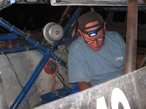 1990 Ford Bronco - man in a cap and headlamp reaches into the blue tube-frame chassis of an off-road race car during a night repair.