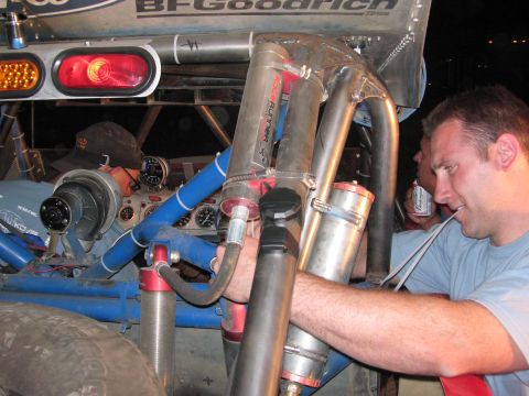 1990 Ford Bronco - man works on the rear suspension and exposed chassis of a BFGoodrich-decaled off-road race truck under night lighting.