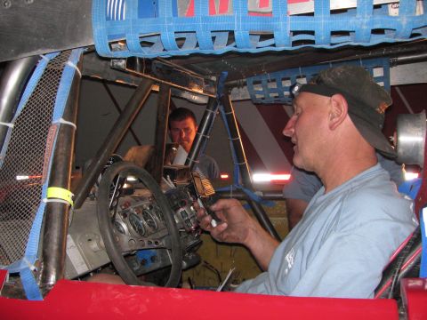 1990 Ford Bronco - Two men work inside an off-road race car cockpit, one handling electronics near the removable steering wheel at night.