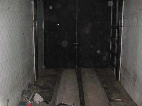 1990 Ford Bronco - Dimly lit interior of an empty enclosed cargo trailer with quilted aluminum walls, floor ramp rails and tools on the floor.