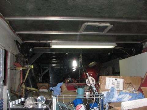 1990 Ford Bronco - dim interior of an enclosed cargo trailer packed with boxes, tools, and equipment under a single fluorescent light strip.