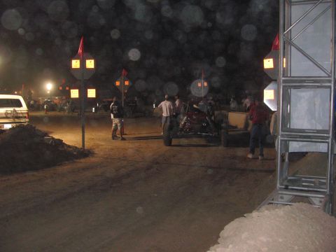 1990 Ford Bronco - nighttime desert race staging area with lit course-marker arrow signs, red flags, spectators, and an off-road buggy on dirt.