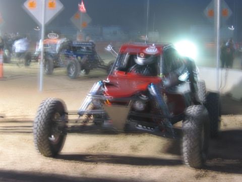 1990 Ford Bronco - red tube-frame off-road race buggy with large knobby tires sits on a dirt course at night with other buggies behind it.