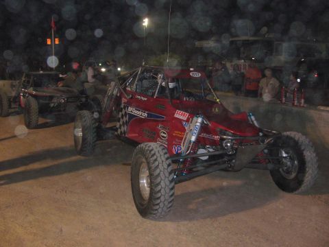 1990 Ford Bronco - red tube-frame off-road race buggy with sponsor decals and knobby tires sits on a dirt course at night near spectators.