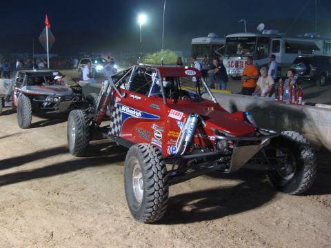 1990 Ford Bronco - red sponsor-decaled off-road buggy and a second buggy are staged on a dirt course at night near a parked bus and crowd.