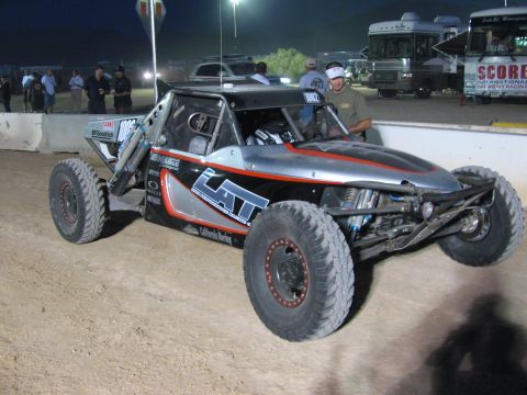 1990 Ford Bronco - black-and-silver tube-frame off-road race buggy with bypass shocks and knobby tires sits staged on a dirt course at night.