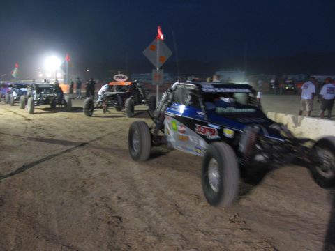 1990 Ford Bronco - blue off-road race buggy leads a line of buggies staged on a dirt lane at dusk beside a lit arrow course-marker sign.