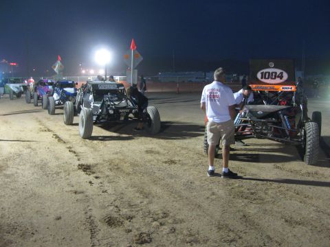 1990 Ford Bronco - Several off-road race buggies line up on a dirt staging lane at dusk while an official in a white shirt walks alongside.