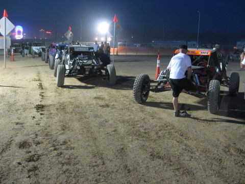 1990 Ford Bronco - row of off-road race buggies sits staged on dirt at night while a crew member pushes one buggy into the lineup.