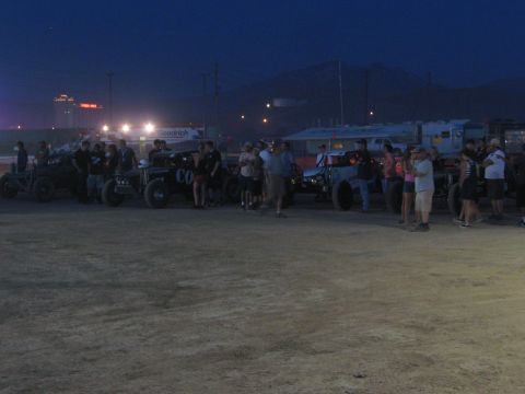 1990 Ford Bronco - Off-road race trucks and buggies gather with spectators on a dusty lot at dusk, mountains and stadium lights behind.